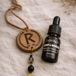 Op de afbeelding: Een houten auto-geurverspreider met de letter "R" gegraveerd, naast een kleine fles etherische olie. De fles is gelabeld "Deep & Woody" en bevat 10 ml olie. De geurverspreider heeft een leren koord en decoratieve kralen.