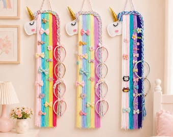 Op de afbeelding: Drie wandhangende eenhoorn-haaraccessoire-organizers gemaakt van kleurrijke garenstrengen. Elk exemplaar heeft een witte eenhoornkop met een gouden hoorn, een gevlochten manen en diverse haarspeldjes, strikjes en haarbanden aan het garen.