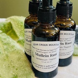 May include: Three amber glass dropper bottles with black caps are arranged on a white surface with a green leaf. The bottles have white labels with black text that reads "Mullein Root" and "Bear Creek Mountain".