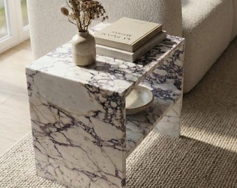 May include: A marble side table with a white and purple veined pattern. The table has a lower shelf and is topped with books, a small vase, and dried flowers. The table is next to a beige sofa and a beige rug.