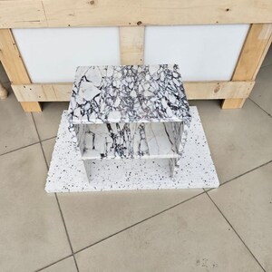 May include: A small, two-tiered marble shelf with a white and black veined pattern. The shelf is placed on a white speckled surface. The background includes a wooden crate and a tiled floor.