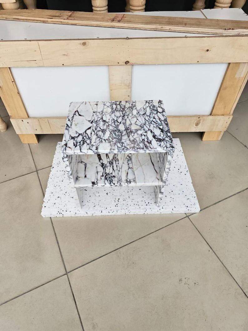 May include: A small, two-tiered marble shelf with a white and black veined pattern. The shelf is placed on a white speckled surface. The background includes a wooden crate and a tiled floor.