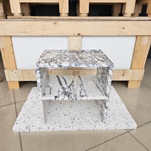 May include: A small, open-shelved side table with a white and gray marble pattern. The table is rectangular with a shelf in the middle. It sits on a white base with black speckles. The background includes wooden furniture.