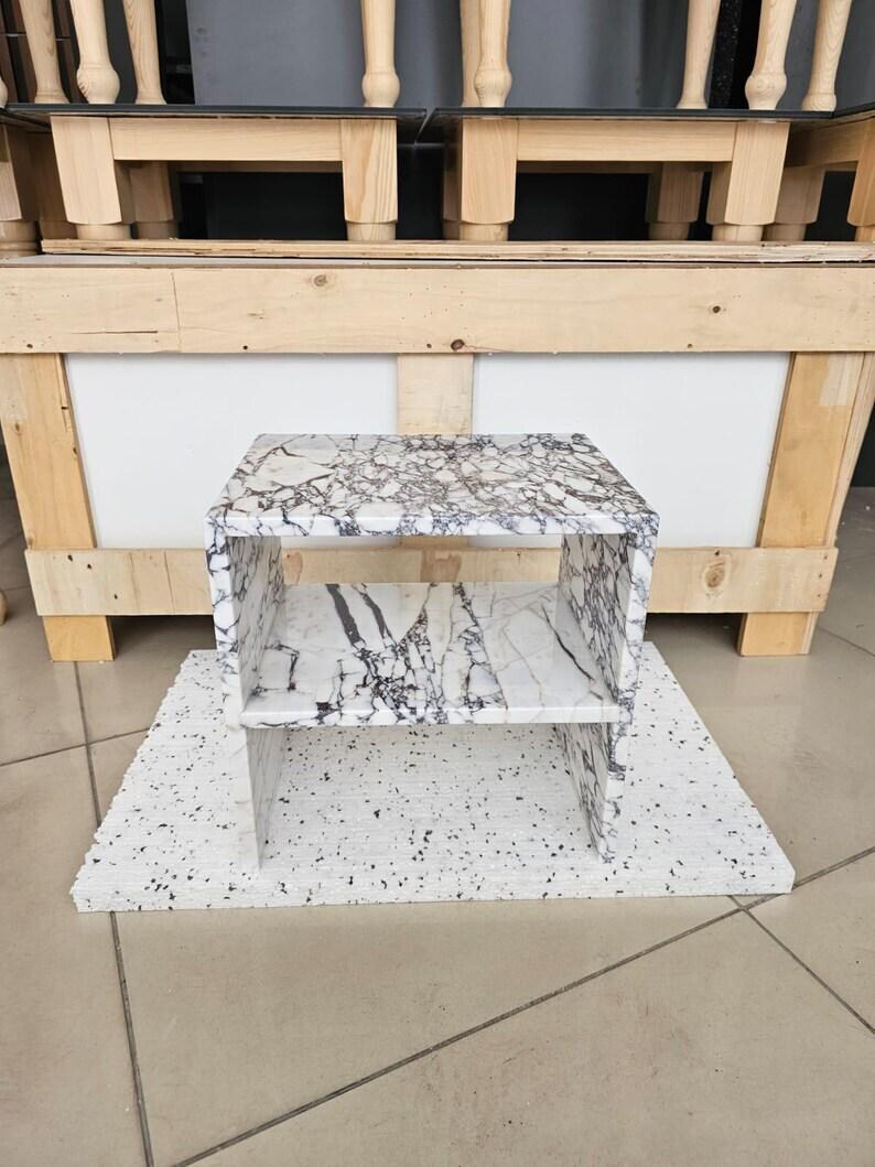 May include: A small, open-shelved side table with a white and gray marble pattern. The table is rectangular with a shelf in the middle. It sits on a white base with black speckles. The background includes wooden furniture.