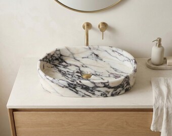 May include: Oval marble sink with black veining, set on a light-colored countertop. Gold-toned faucet and drain. A white soap dispenser and a bar of soap are on the right. A linen towel hangs below.