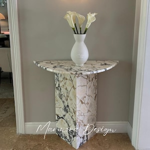 Puede incluir: Una consola de mármol con una tapa semicircular y una base rectangular. La mesa presenta un patrón veteado blanco y negro. Un jarrón blanco con diseño de rostro contiene calas blancas. La mesa está contra una pared gris.