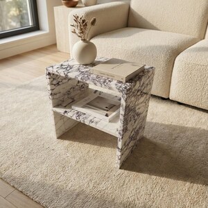 May include: A marble side table with a white and purple veined pattern. The table has a shelf and holds a book and a small vase with dried flowers. The table is on a beige rug.