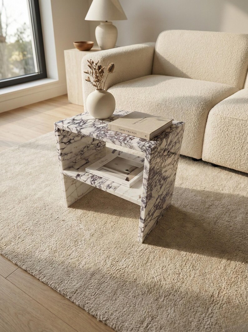 May include: A marble side table with a white and purple veined pattern. The table has a shelf and holds a book and a small vase with dried flowers. The table is on a beige rug.