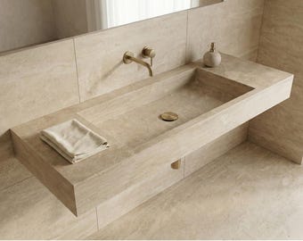 May include: A modern bathroom sink made of light beige stone. The sink has a rectangular basin, a gold faucet, and a matching drain cover. A folded beige towel rests on the counter. A matching soap dispenser is also present.