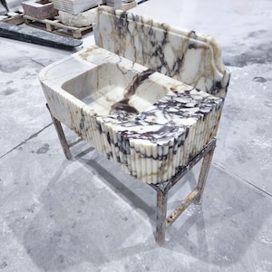May include: A marble bathroom sink with a rectangular basin and a decorative, fluted exterior. The sink is supported by a metal stand. The marble features a white and brown veined pattern. Another marble piece is behind the sink.