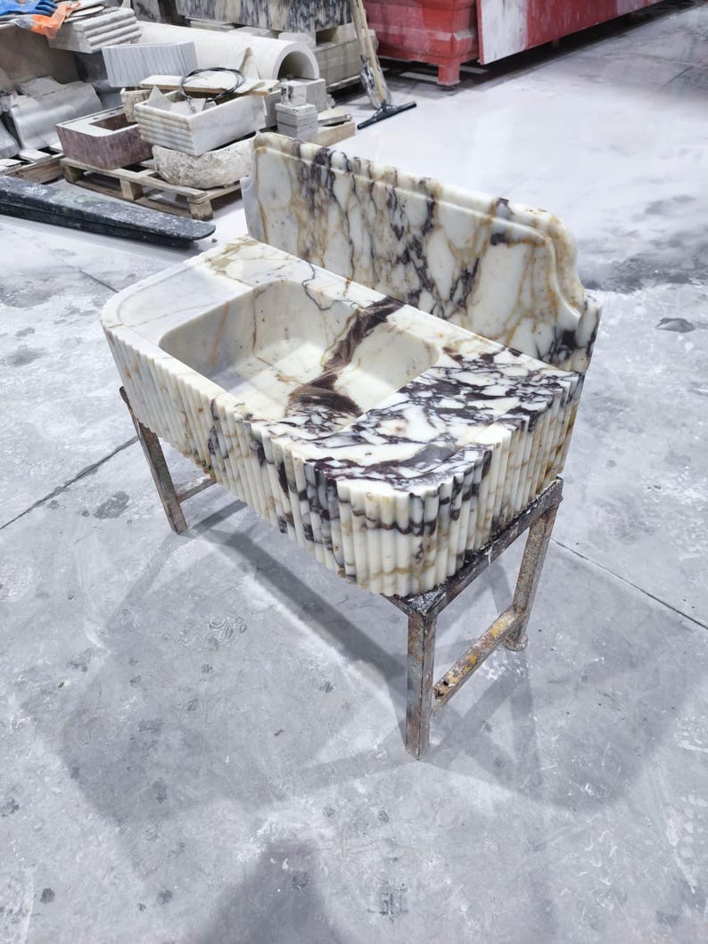 May include: A marble bathroom sink with a rectangular basin and a decorative, fluted exterior. The sink is supported by a metal stand. The marble features a white and brown veined pattern. Another marble piece is behind the sink.