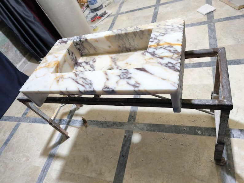 Puede incluir: Un lavabo rectangular de m&aacute;rmol con un patr&oacute;n veteado blanco, morado y dorado. El lavabo est&aacute; sostenido por un marco de metal oscuro. El lavabo es probablemente para un ba&ntilde;o o una cocina.