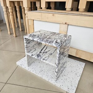 May include: A small, rectangular marble side table with a white and gray veined pattern. The table has two shelves and sits on a white speckled base. The background shows other wooden furniture.