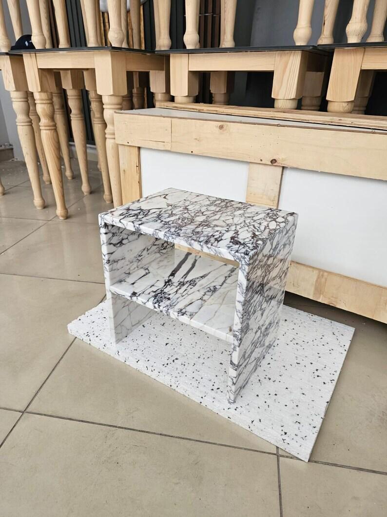 May include: A small, rectangular marble side table with a white and gray veined pattern. The table has two shelves and sits on a white speckled base. The background shows other wooden furniture.