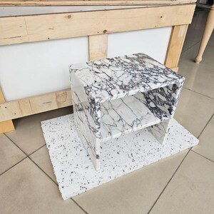 May include: A white and black marble-patterned side table with a shelf. The table is rectangular and sits on a white, speckled surface. The table is made of a solid material and has a modern design.