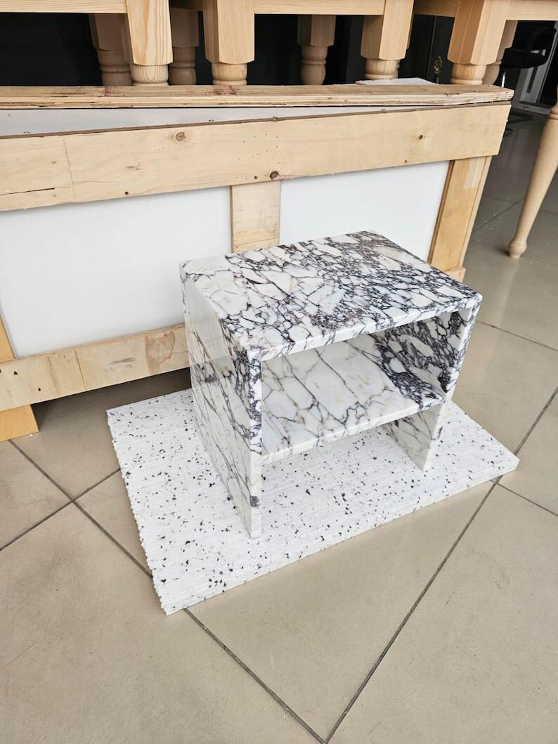 May include: A white and black marble-patterned side table with a shelf. The table is rectangular and sits on a white, speckled surface. The table is made of a solid material and has a modern design.