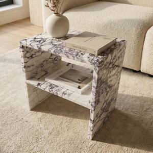 May include: A marble side table with a white and purple veined pattern. The table has two shelves and holds a book and a magazine. A white vase with dried flowers sits on top. The table is on a beige rug.