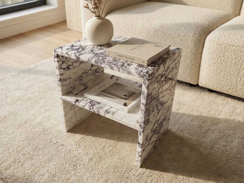 May include: A marble side table with a white and purple veined pattern. The table has two shelves and holds a book and a magazine. A white vase with dried flowers sits on top. The table is on a beige rug.