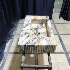 Puede incluir: Lavabo rectangular de m&aacute;rmol con dise&ntilde;o de borde acanalado. La cubeta del lavabo es blanca con vetas grises y doradas. El lavabo est&aacute; montado sobre un soporte de metal oscuro. Es un accesorio de ba&ntilde;o.