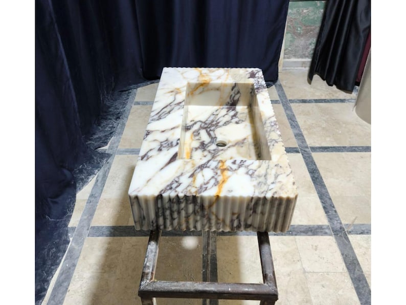 Puede incluir: Lavabo rectangular de m&aacute;rmol con dise&ntilde;o de borde acanalado. La cubeta del lavabo es blanca con vetas grises y doradas. El lavabo est&aacute; montado sobre un soporte de metal oscuro. Es un accesorio de ba&ntilde;o.