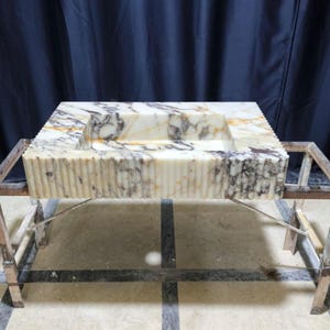 Puede incluir: Un lavabo rectangular de m&aacute;rmol con una pila tallada, sostenido por un marco de metal. El lavabo presenta un patr&oacute;n veteado crema y gris con detalles acanalados verticales en los lados. El marco de metal tiene una apariencia r&uacute;stica e industrial.