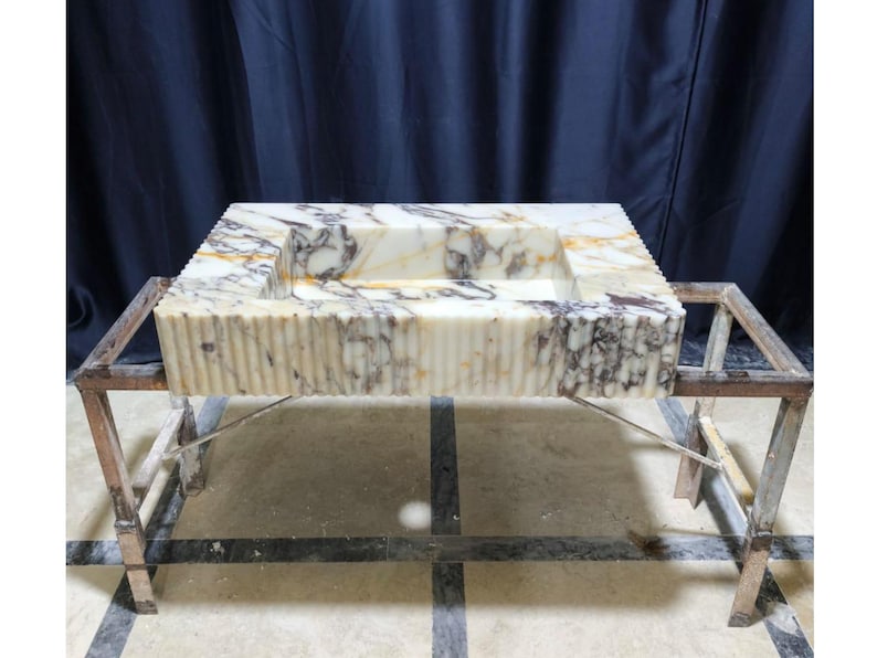 Puede incluir: Un lavabo rectangular de m&aacute;rmol con una pila tallada, sostenido por un marco de metal. El lavabo presenta un patr&oacute;n veteado crema y gris con detalles acanalados verticales en los lados. El marco de metal tiene una apariencia r&uacute;stica e industrial.