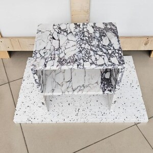 May include: A small, rectangular marble table with a white and black veined pattern. The table has a shelf and is placed on a white, speckled surface. The table is approximately 18 inches long, 12 inches wide, and 12 inches tall.
