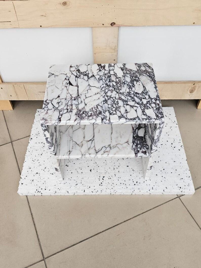 May include: A small, rectangular marble table with a white and black veined pattern. The table has a shelf and is placed on a white, speckled surface. The table is approximately 18 inches long, 12 inches wide, and 12 inches tall.
