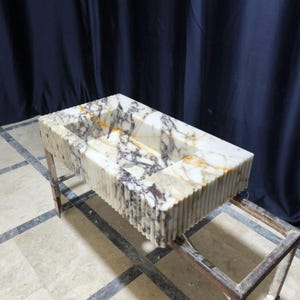 Puede incluir: Lavabo rectangular de m&aacute;rmol con exterior estriado. El lavabo presenta una paleta de colores naturales de blanco, marr&oacute;n y gris. Est&aacute; soportado por un marco met&aacute;lico, mostrando una mezcla de texturas y materiales.