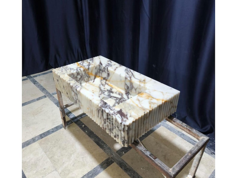 Puede incluir: Lavabo rectangular de m&aacute;rmol con exterior estriado. El lavabo presenta una paleta de colores naturales de blanco, marr&oacute;n y gris. Est&aacute; soportado por un marco met&aacute;lico, mostrando una mezcla de texturas y materiales.
