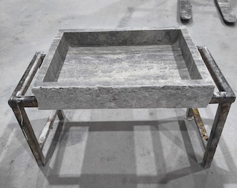 May include: A large, square, gray stone basin with a recessed interior sits on a metal stand. The basin has a textured surface and a rectangular shape. The metal stand has a weathered appearance.