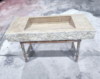 May include: A rectangular stone sink with a built-in basin. The sink is light beige with a rough, textured surface. It rests on a rusty metal stand with four legs. The sink is likely for a bathroom or kitchen.