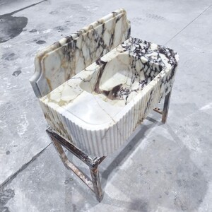 May include: A marble sink with a rectangular basin and a matching backsplash. The sink has a white and brown veined pattern and is supported by a metal stand. The sink's front has a fluted design.