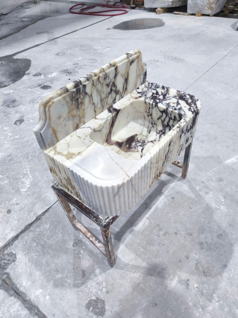 May include: A marble sink with a rectangular basin and a matching backsplash. The sink has a white and brown veined pattern and is supported by a metal stand. The sink's front has a fluted design.