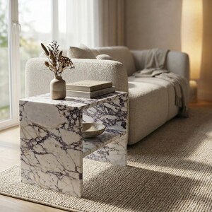 May include: A marble side table with a shelf, featuring a white and purple veined pattern. The table holds a small vase with dried flowers, books, and a bowl. The setting includes a beige sofa, rug, and natural light.