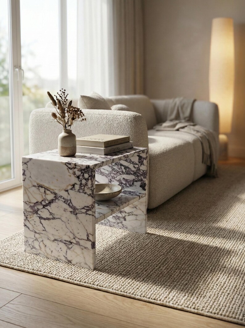 May include: A marble side table with a shelf, featuring a white and purple veined pattern. The table holds a small vase with dried flowers, books, and a bowl. The setting includes a beige sofa, rug, and natural light.