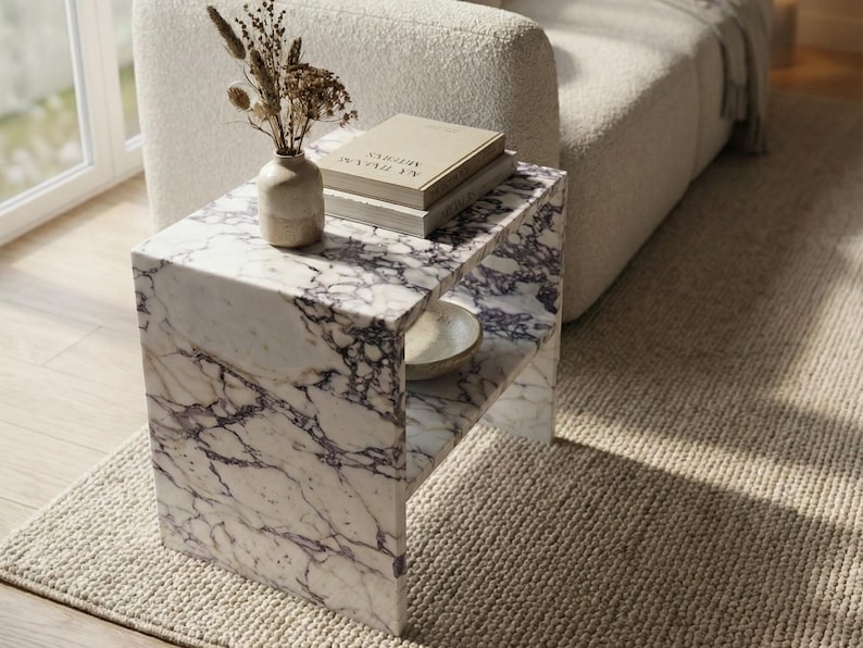 May include: A marble side table with a white and purple veined pattern. The table has a lower shelf and is topped with books, a small vase, and dried flowers. The table is next to a beige sofa and a beige rug.