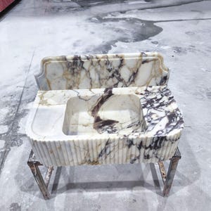 May include: A marble sink with a rectangular basin and a decorative backsplash. The sink features a white and brown veined pattern. The front of the sink has vertical ribbed detailing. It is supported by a metal stand.