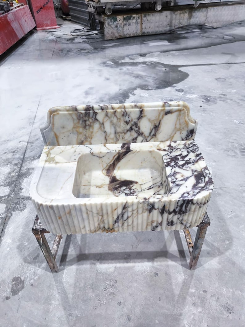 May include: A marble sink with a rectangular basin and a decorative backsplash. The sink features a white and brown veined pattern. The front of the sink has vertical ribbed detailing. It is supported by a metal stand.