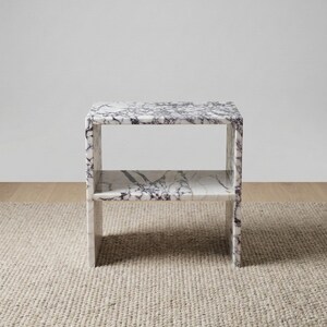 May include: A marble side table with a shelf. The table is white with dark gray veining. It has a rectangular shape and is placed on a beige rug. The table is approximately 24 inches tall, 18 inches wide, and 12 inches deep.