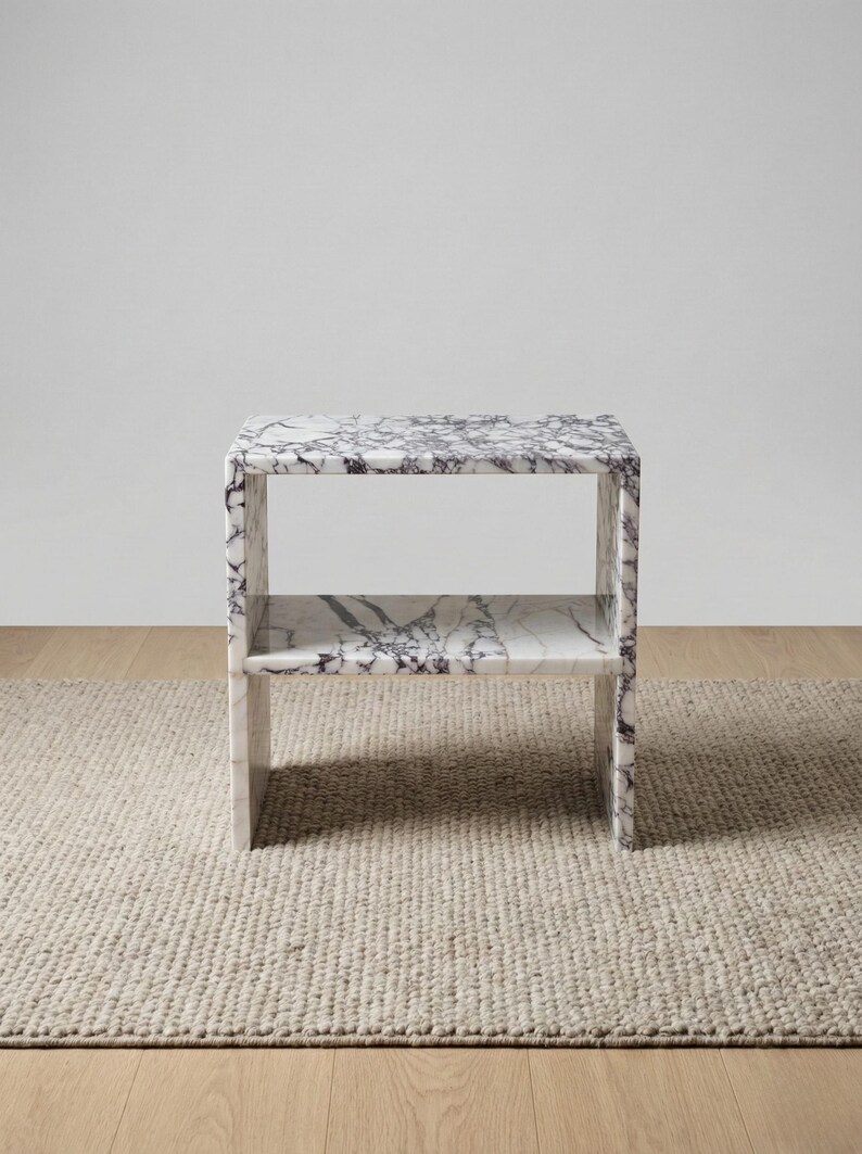 May include: A marble side table with a shelf. The table is white with dark gray veining. It has a rectangular shape and is placed on a beige rug. The table is approximately 24 inches tall, 18 inches wide, and 12 inches deep.