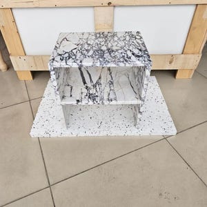 May include: A small, two-tiered shelf unit with a white and black marble pattern. The unit is made of a solid material and sits on a matching base. The shelf is approximately 12 inches wide and 10 inches tall.