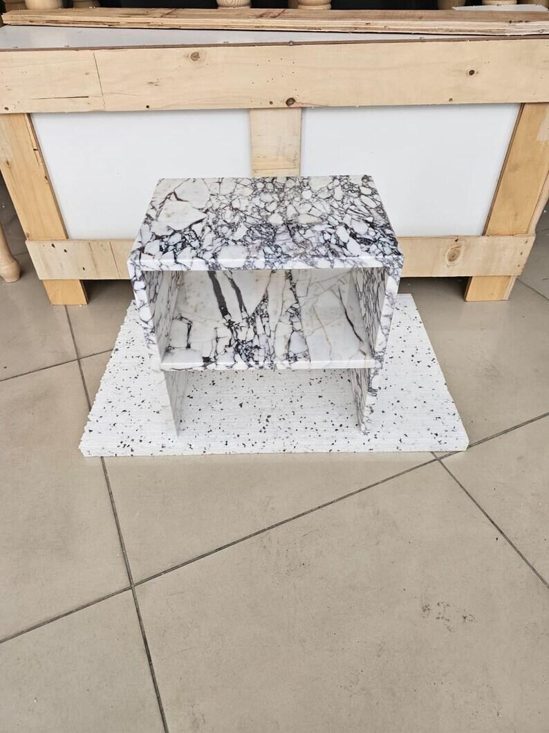 May include: A small, two-tiered shelf unit with a white and black marble pattern. The unit is made of a solid material and sits on a matching base. The shelf is approximately 12 inches wide and 10 inches tall.