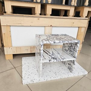 May include: A small, marble-look side table with a shelf. The table has a white base with black veining and is set on a white, speckled base. The background shows wooden furniture legs and a wooden structure.