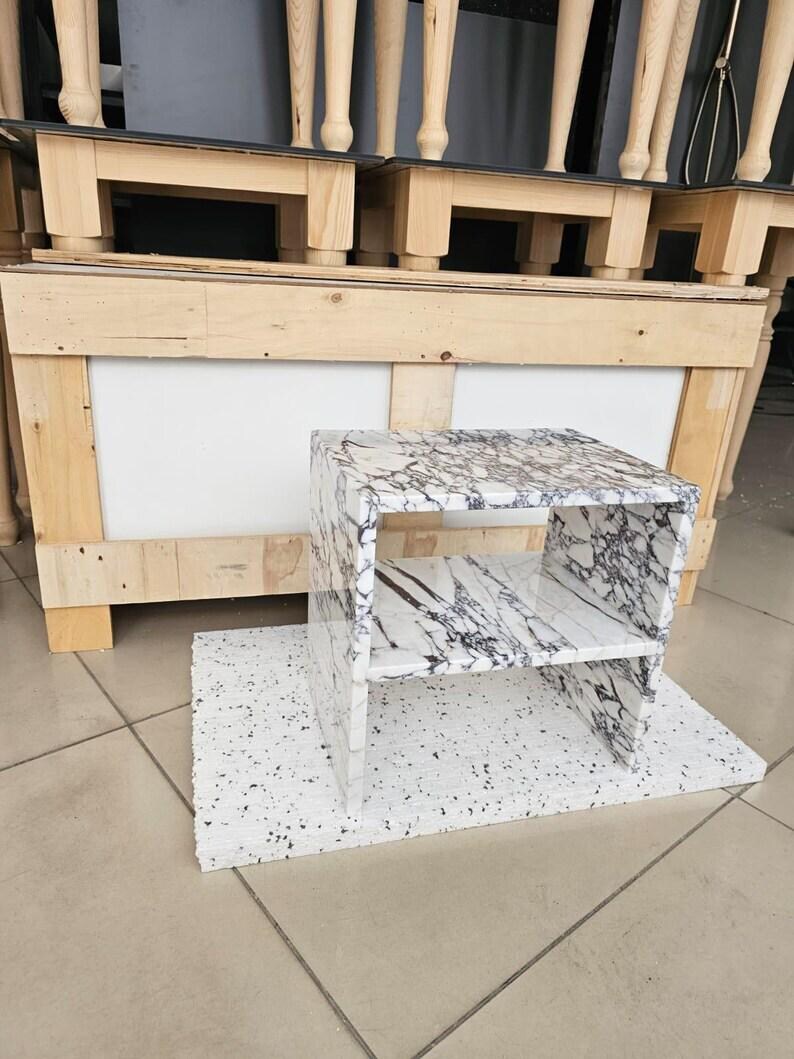 May include: A small, marble-look side table with a shelf. The table has a white base with black veining and is set on a white, speckled base. The background shows wooden furniture legs and a wooden structure.