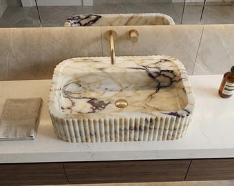 Peut inclure: Un évier rectangulaire en marbre avec un extérieur cannelé et un robinet doré. L'évier présente un motif veiné crème et violet. Une serviette pliée et une bouteille sont sur le comptoir.