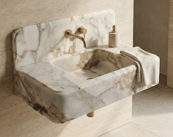 May include: A wall-mounted marble sink with a built-in backsplash. The sink features a gold faucet and soap dispenser. A folded towel rests on the edge. The marble has a white and brown veined pattern.