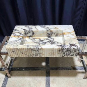 Puede incluir: Fregadero rectangular de m&aacute;rmol con una cubeta tallada. El fregadero presenta un patr&oacute;n veteado crema y gris y un dise&ntilde;o de borde acanalado. Se exhibe sobre un soporte met&aacute;lico.