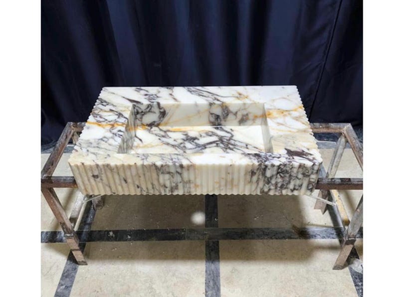 Puede incluir: Fregadero rectangular de m&aacute;rmol con una cubeta tallada. El fregadero presenta un patr&oacute;n veteado crema y gris y un dise&ntilde;o de borde acanalado. Se exhibe sobre un soporte met&aacute;lico.