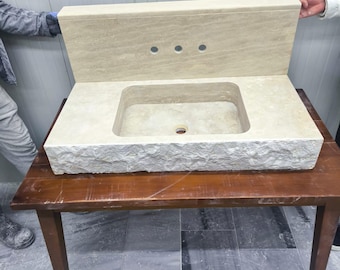 May include: A beige stone bathroom sink with a rectangular basin and a backsplash featuring three faucet holes. The sink has a rough-hewn edge and is displayed on a wooden table. The overall design suggests a natural, rustic aesthetic.
